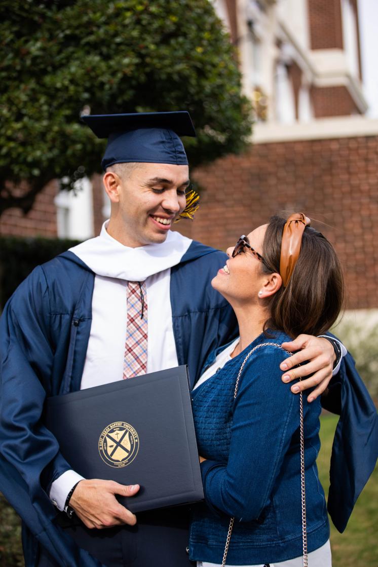 A man in his graduation regalia and a woman looking at each other smiling outside