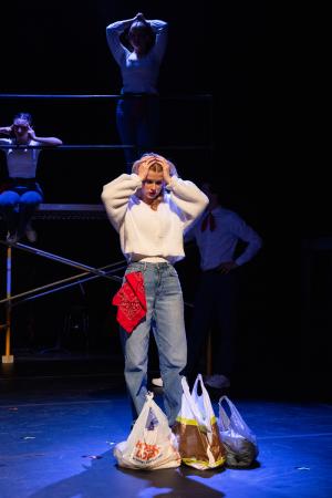 A woman holding her head with three bags in front of her feet in Working: The Musical Theatre production.