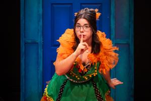 Woman looking at camera with finger pressed up against lips in front of blue door during Cagebirds Theatre Production