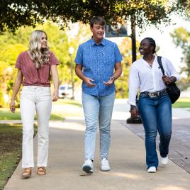 Two females and one male walking down a sidewalk talking outside on a sunny day.