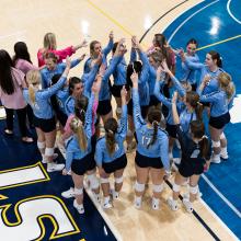 Overhead of group of volleyball girls on the court in a circle with their arms in the air in the middle of the circle