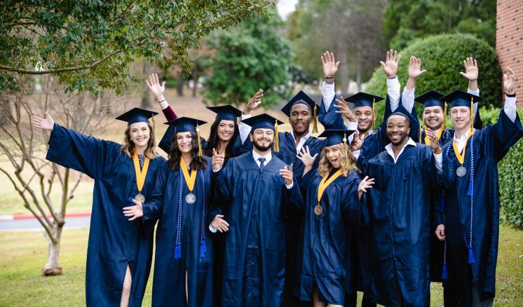 Group of graduates in their caps and gowns smiling at the camera with their hands up