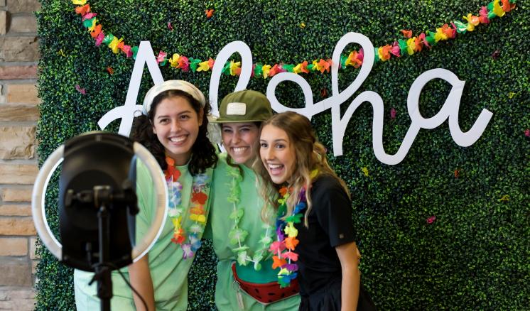 Three women taking pictures in front of a ring light with a plant backdrop with the word aloha in the back
