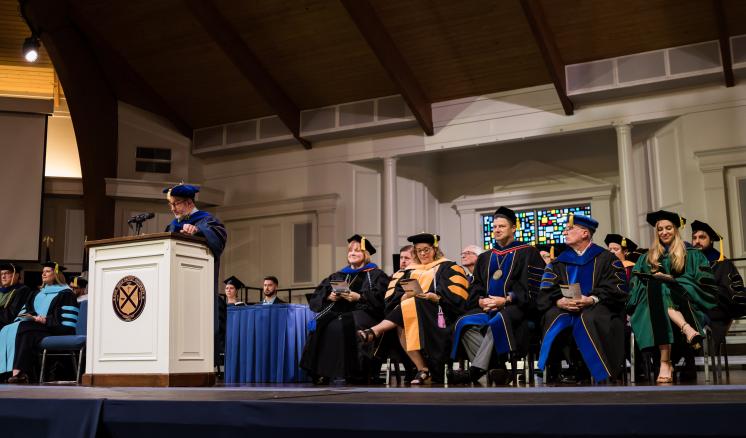 Man speaking in graduation regalia as well as others in graduation regalia in the background