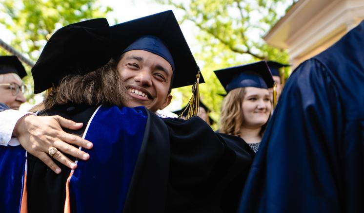 Student in graduation regalia hugging a professor in graduation regalia