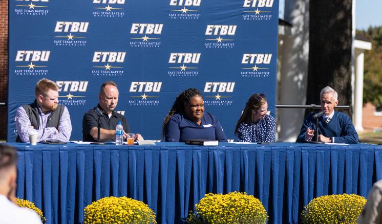 Men and women sitting at a table with a blue tablecloth in front of a blue ETBU backdrop outside