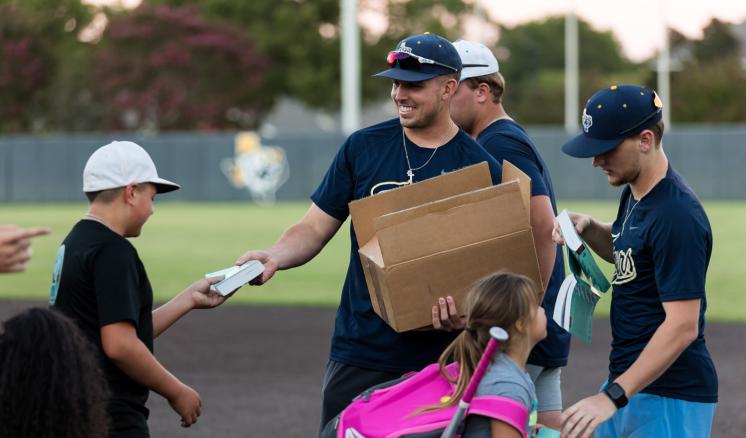 Two men handing out books to little kids from a box on the baseball field