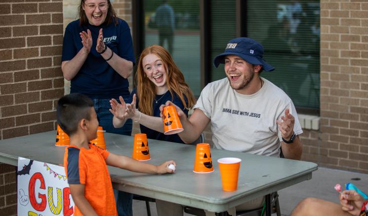 A man and woman sitting at a table with pumpkin cups smiling at a little boy with a woman clapping her hands in the background