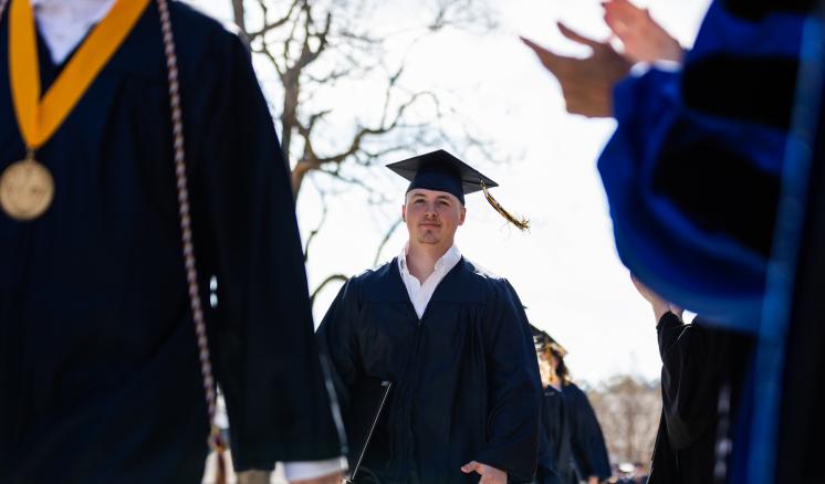 Male student in cap and gown walking outdoors with diploma in his hand