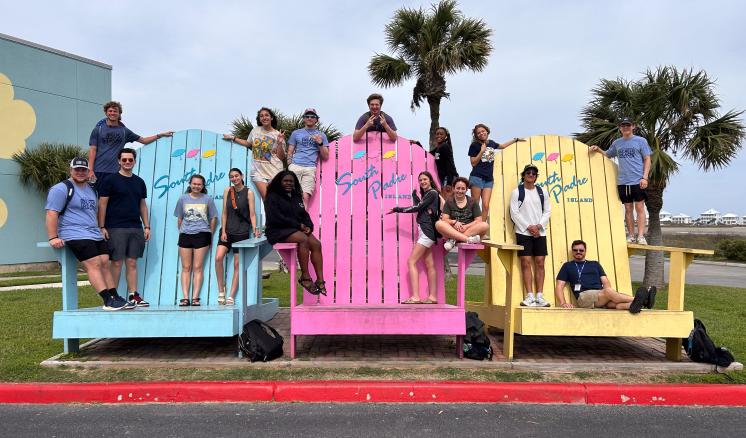 A group of individuals sit and stand on three oversized wooden chairs next to the beach at South Padre Island, Texas