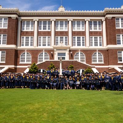 A group of college graduates in navy caps and gowns hold their degrees in front of a red brick building