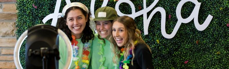 Three women taking pictures in front of a ring light with a plant backdrop with the word aloha in the back