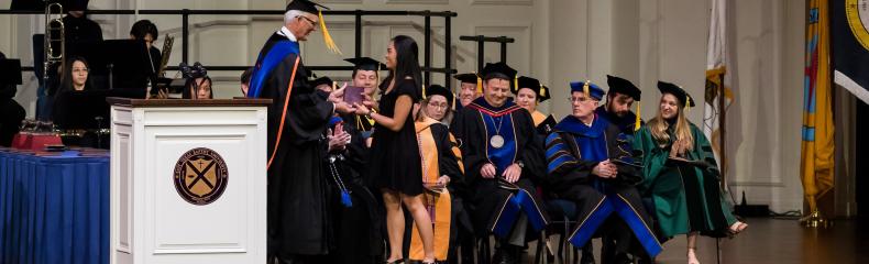 Man in graduation regalia giving a woman a plaque with others in the graduation regalia in the background