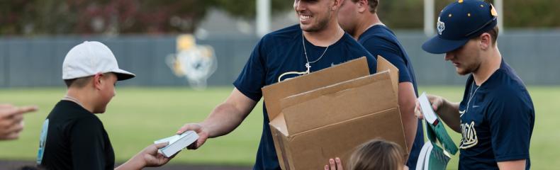 Two men handing out books to little kids from a box on the baseball field