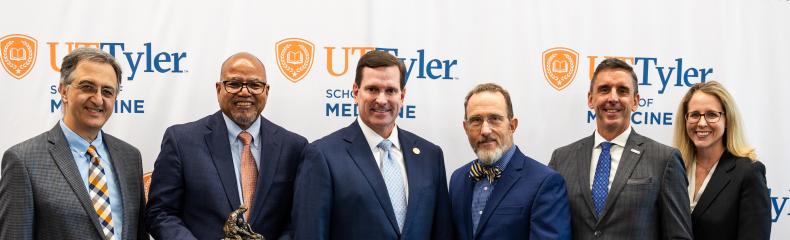 Group of five males and one female smiling at camera in front of a white UT Tyler backdrop