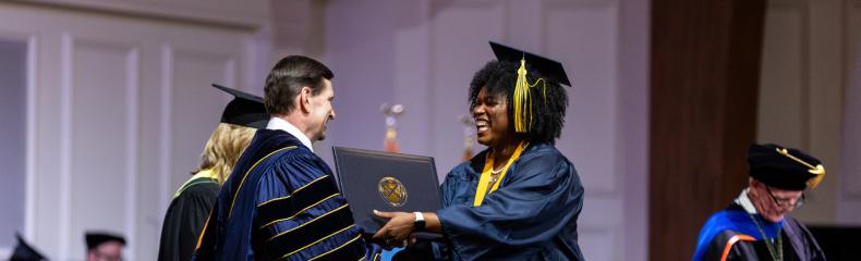 During a commencement ceremony, a woman in a navy cap and gown on the right receive her diploma from a man in navy academic regalia on the left.