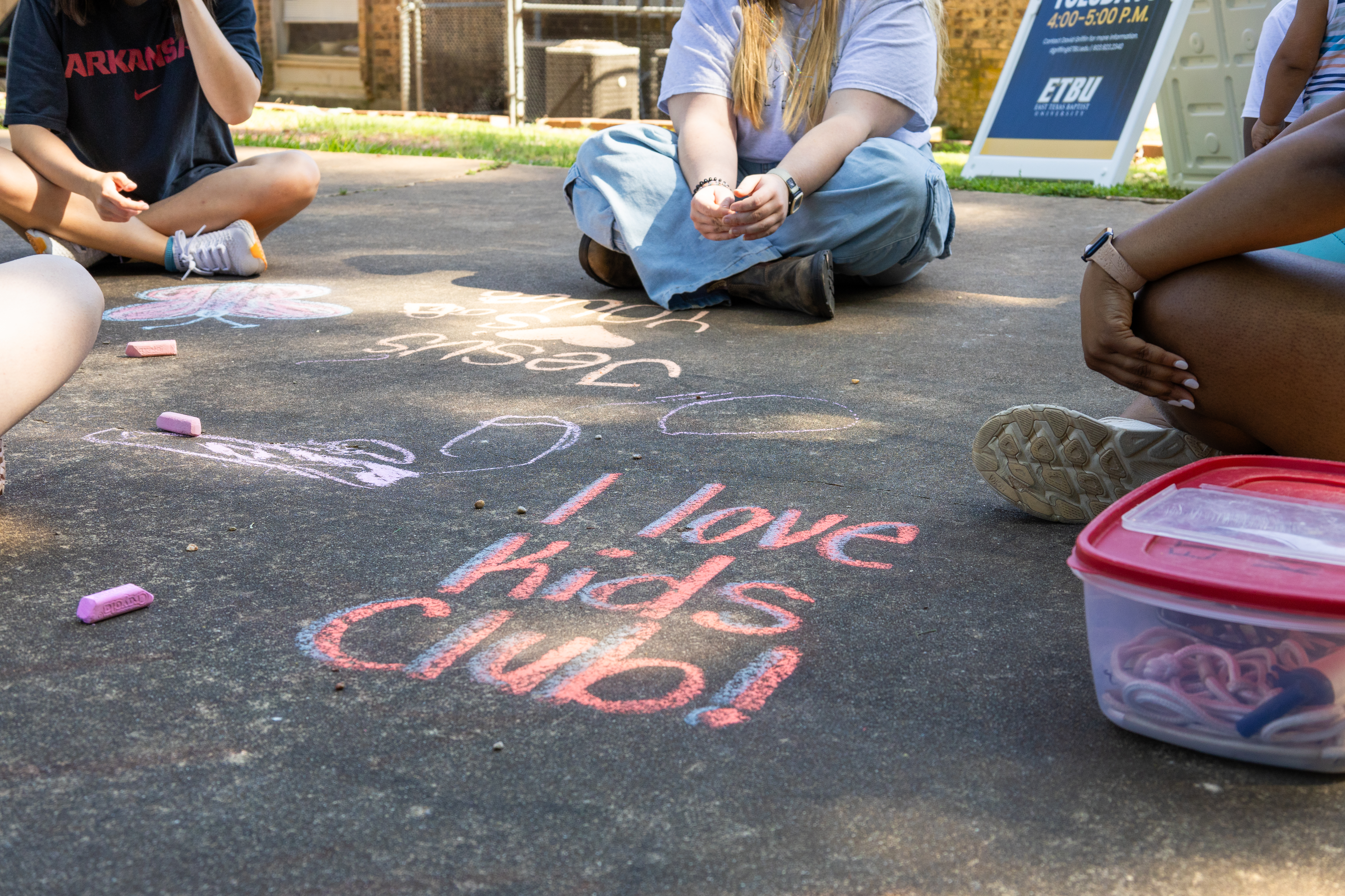 People sitting in a circle on pavement with chalk drawings and the writing "I love Kids Club!".