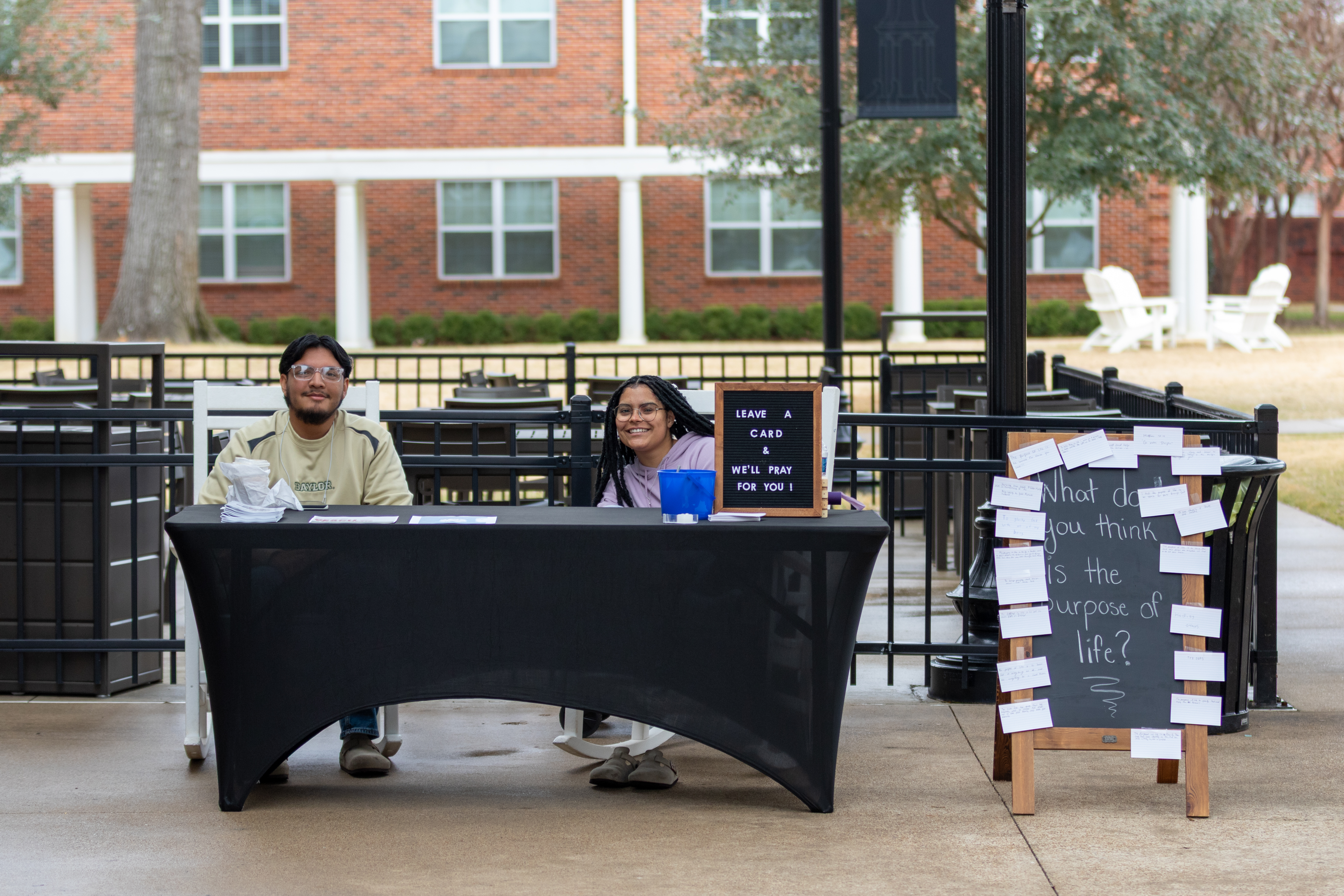 Two people seated at a table outdoors with a prayer invitation sign and a chalkboard asking about the purpose of life.