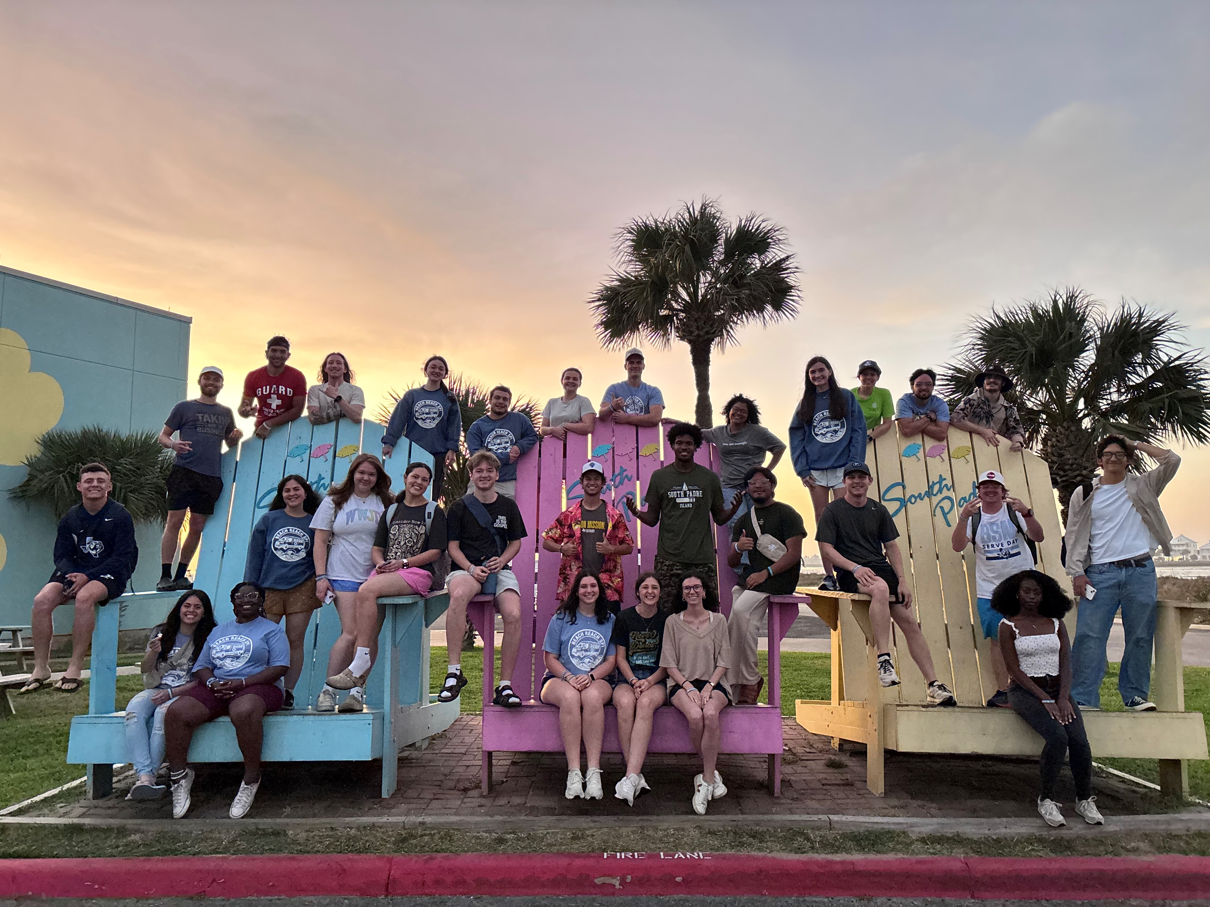 A group of students sitting and standing on large pastel-colored Adirondack chairs in front of a sunset.