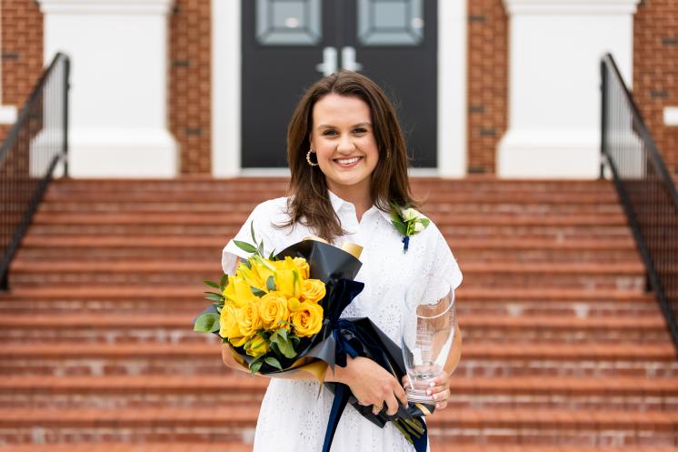 Woman smiling and standing in front of brick steps holding a bouquet of yellow roses and a glass award.