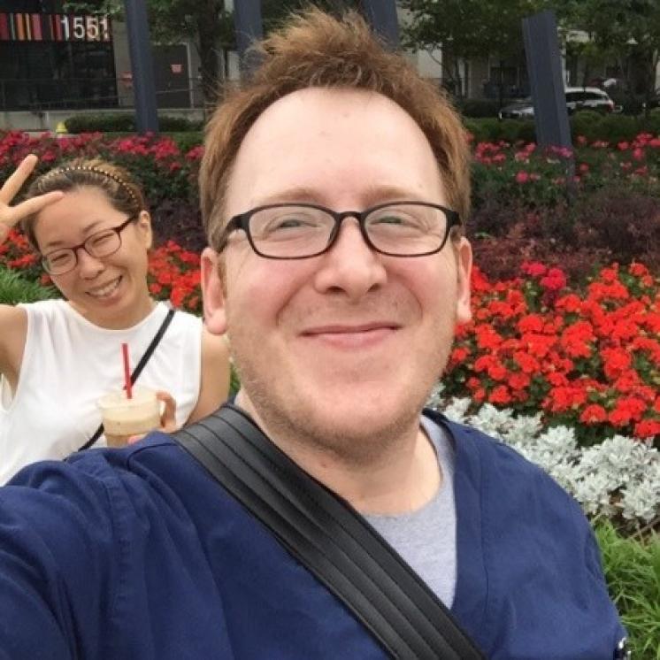 A selfie of a man in scrubs and glasses smiling at a camera with a woman holding up a peace sign on the left in the background.