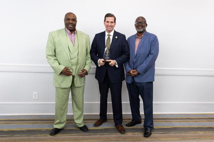 Three men are standing together in a formal setting, with the man in the center holding an award.