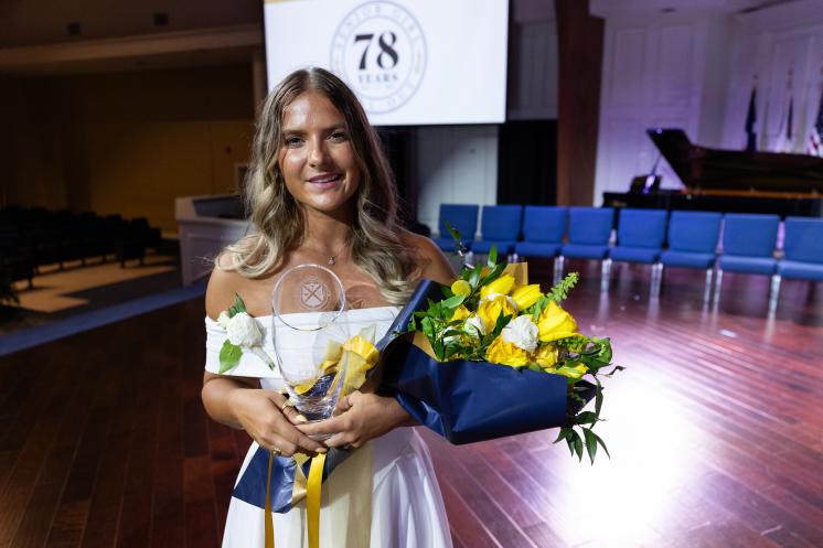 A young woman in a white dress holds a bouquet of yellow flowers and a glass award while standing on a stage during a formal event.
