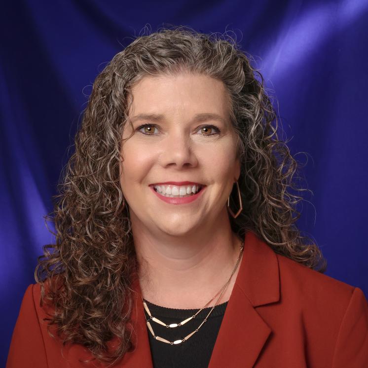 A woman standing smiling with a purple background, gold necklases and a red blazer