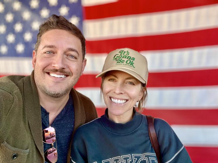 2 people standing smiling in front of an American Flag