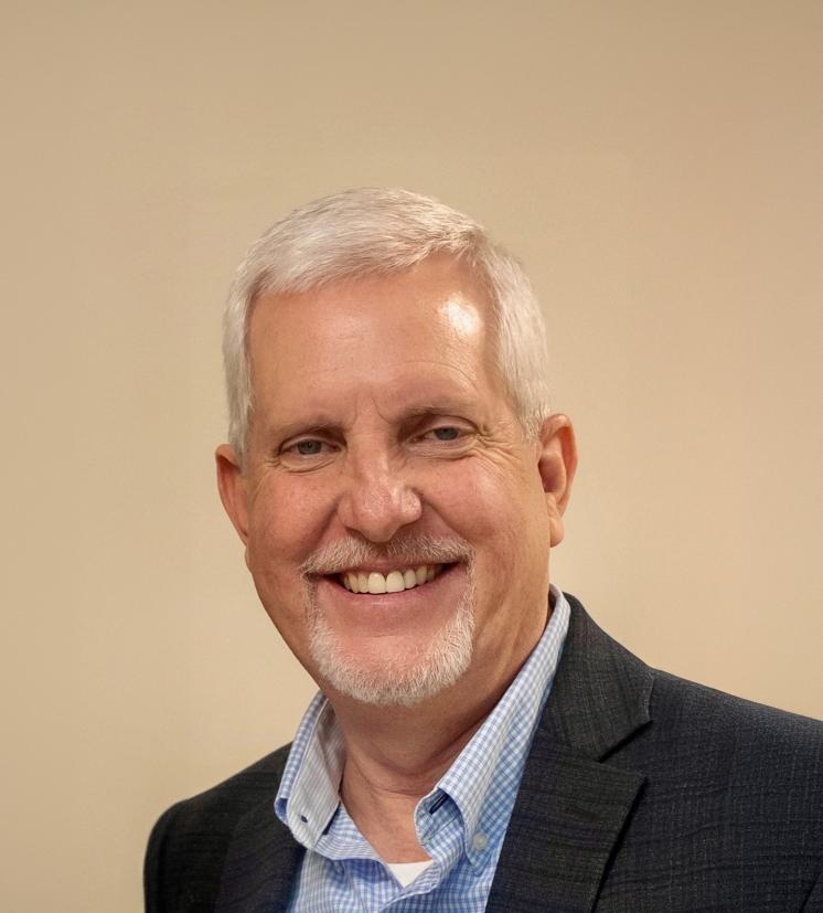 A white man with gray hair smiling in front of a cream wall wearing a blue shirt and gray blazer.