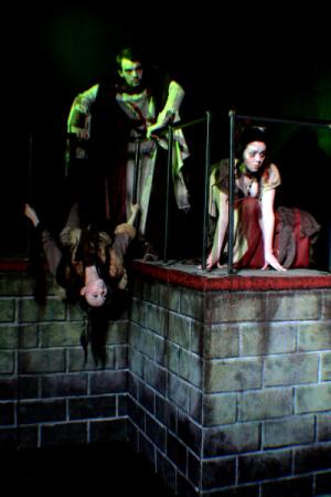 2 women, one hanging upside down on a brick wall while a man stands behind them in dark clothing and lighting