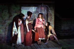 A group of people in ragged and torn clothing standing on a wodden floor with a brick wall behind them, dimly lit, and holding a clear umbrella