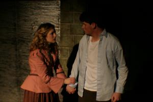 A woman holding a mans hand under dark lighting wearing business casual clothing with a brick wall behind them