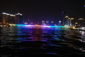 Nighttime cityscape with illuminated buildings and a multicolored bridge reflected on the water.