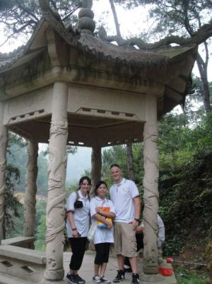 Three people in front of a small pavilion with carved pillars in a wooded area.