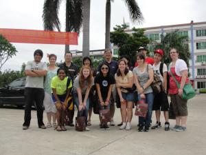 A group of thirteen people stands outside a building with palm trees and a red banner.