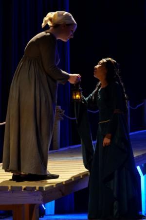 Two women interacting on a dimly lit stage, one handing a lantern to the other.