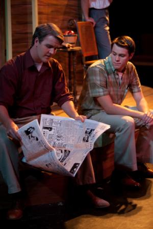 two guys sitting together on a bench, looking down, reading a newspaper in business casual clothing