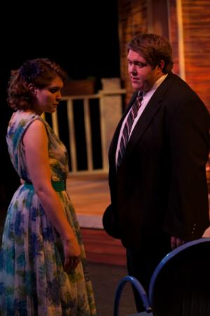 A man and a woman standing on a wooden porch in formal clothing talking to eachother