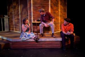 2 men sitting on a prop porch on a stage talking to a woman sitting down in business casual clothing