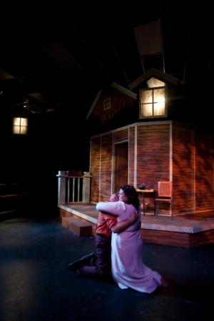 A man hugging a woman on a dimly lit stage with a prop house in the background
