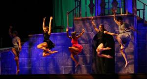 Five dancers in mid-air on a blue-lit stage, with a colorful background.