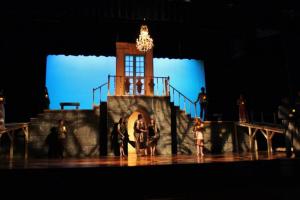 Theatrical stage with period costumed actors on a grand staircase set and blue-lit backdrop.