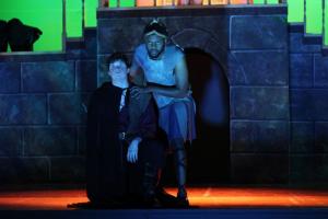 Two performers in medieval costumes on a dimly lit stage with colored lighting.