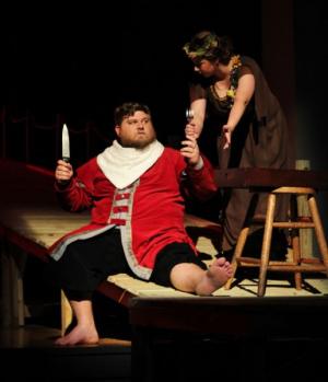 Two actors in a dramatic stage scene, one sitting with a knife and one standing with a small bottle in hand.