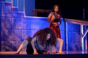 Two performers on stage, one crouching and one standing, with blue-lit stone walls behind them.