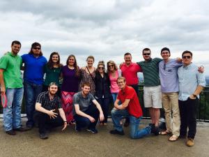 A group of thirteen people posing outdoors on a paved surface with a cloudy sky and landscape in the background.
