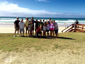 A group of people posing on grass near a beach with ocean waves and a partly cloudy sky.
