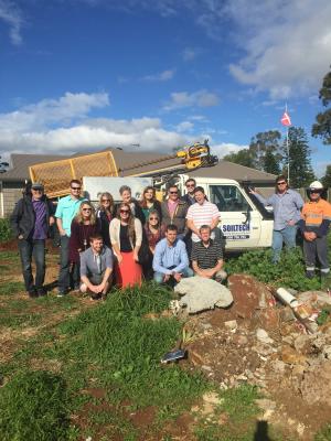 A group of people posing in front of a white truck, with a house and blue sky in the background.