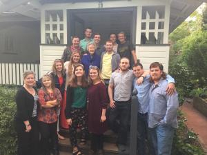 A group of sixteen people posing on a porch in front of a house with greenery around.
